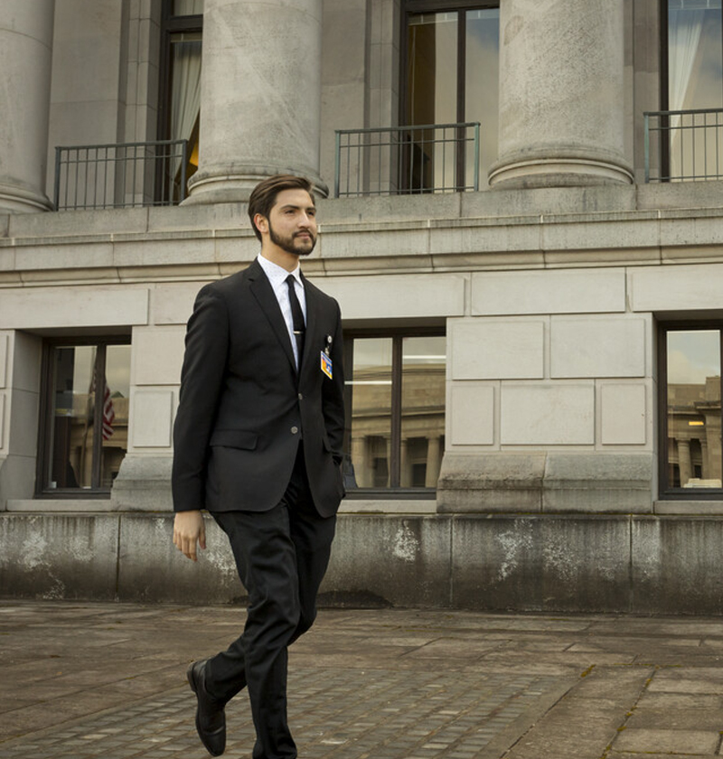Person wearing black suit walks in front of white building with columns.