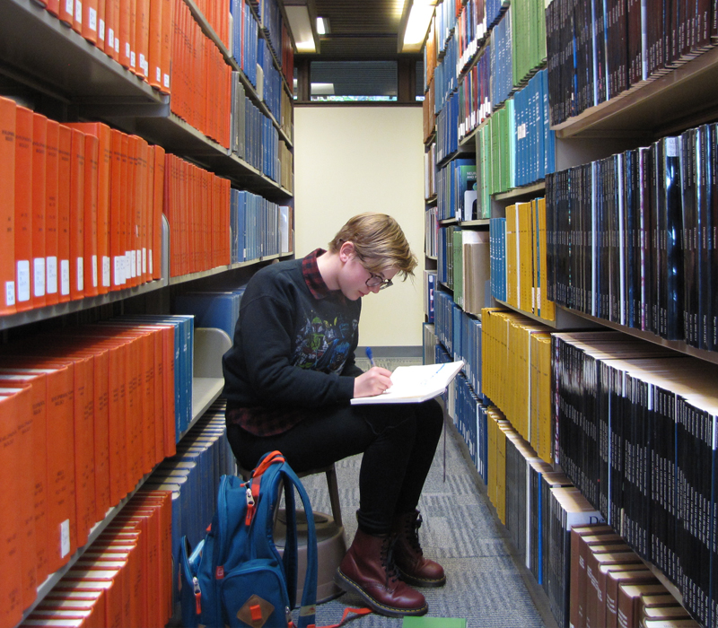 A person sits on a stool in a library between two shelves of books writing in a notebook.