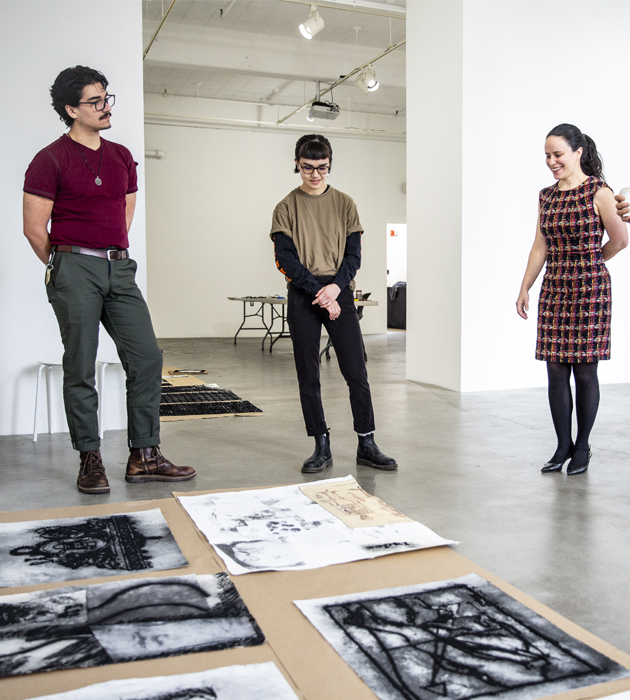 Students and faculty stand in a gallery and look at charcoal drawings on paper on the ground to be hung on the gallery walls.