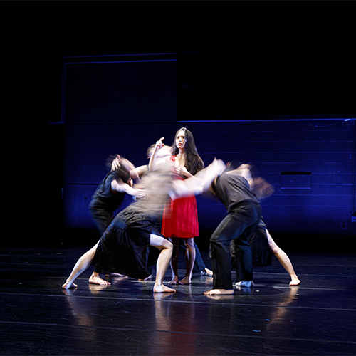 Group of dancers on stage huddle around another dancer in red dress.