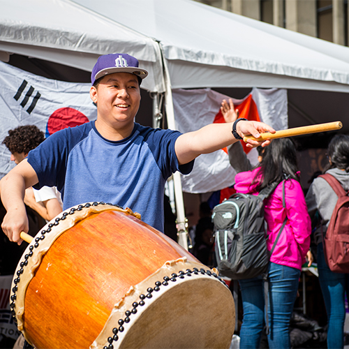 Person with a large wooden drum points a drumstick to the right.