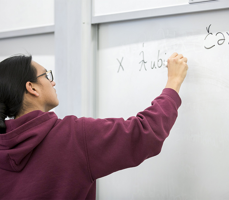 Student in maroon hoodie writing on whiteboard