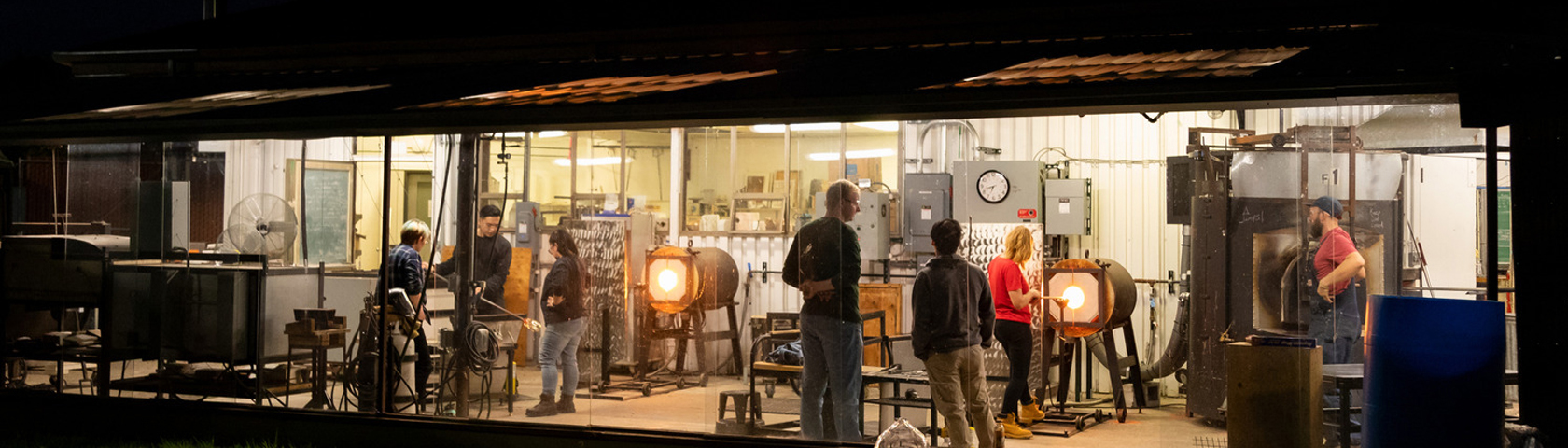 View of glass blowing kilns from outside at night with students blowing glass inside