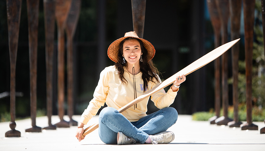 Stephanie Masterman in front of the Burke Museum, holding a cedar oar that she carved.
